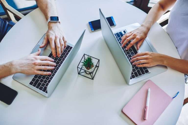 two people typing on their laptops on a white table with a flower in the middle, with a phone also on the table