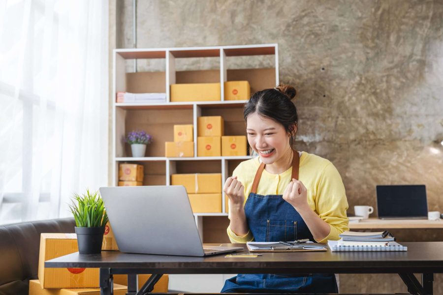 A young Asian woman entrepreneur celebrates excitedly in front of her laptop, wearing a yellow top and blue apron. She's surrounded by shipping boxes and a tidy workspace, embodying the success of mastering WordPress scheduling for her online business.