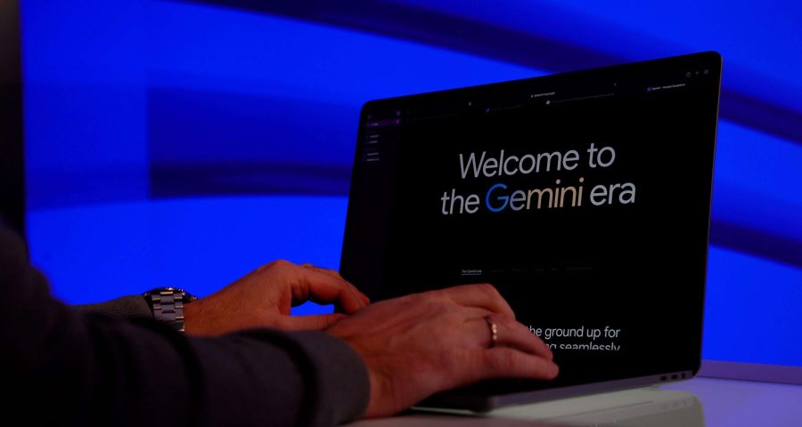A person typing on a laptop that displays a welcome message to the ‘Gemini era’ on its screen, set against a vibrant blue background illuminated by lights.