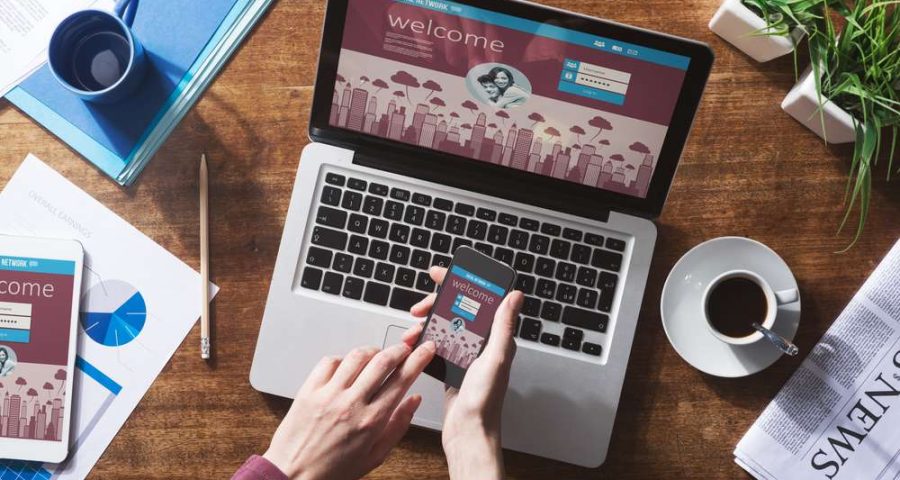 A downward view of a laptop can be seen, with an iPhone and two hands above the keyboard. Two cups of coffee surround the laptop.