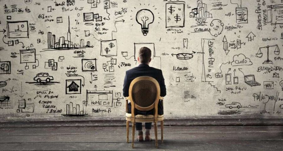 A photograph of a lone man sitting before a wall with his ideas splayed out in front of him in cartoon form.